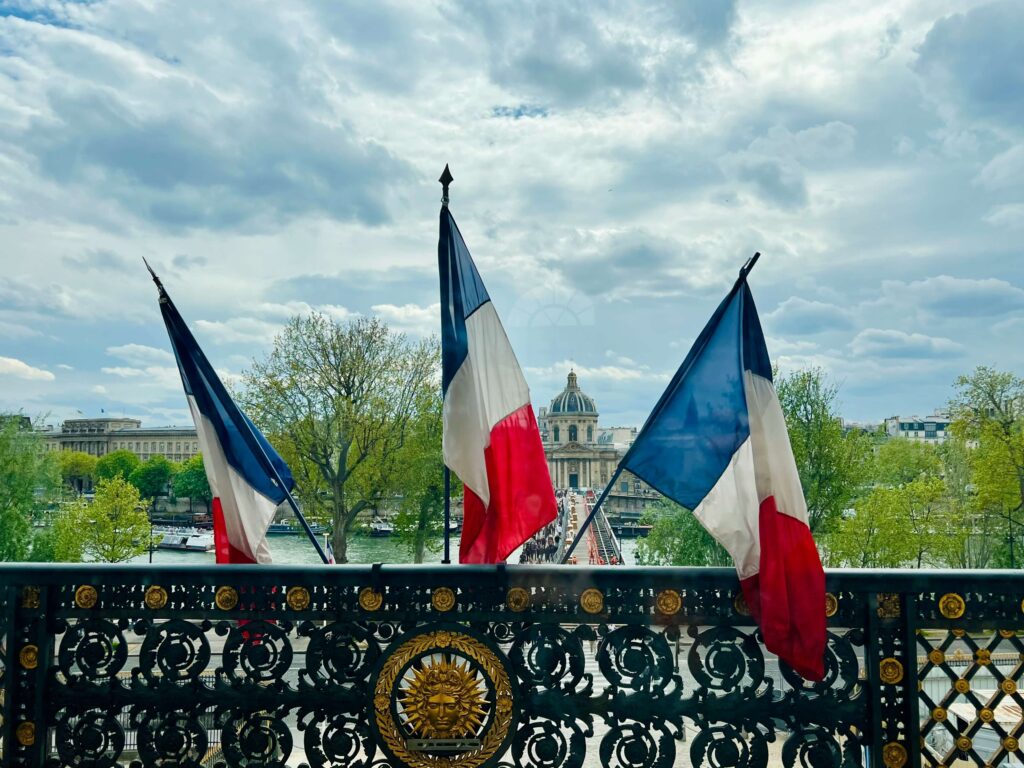 Franse en Europese vlaggen wapperen voor een historisch gebouw met uitzicht over een stad en groene heuvels.